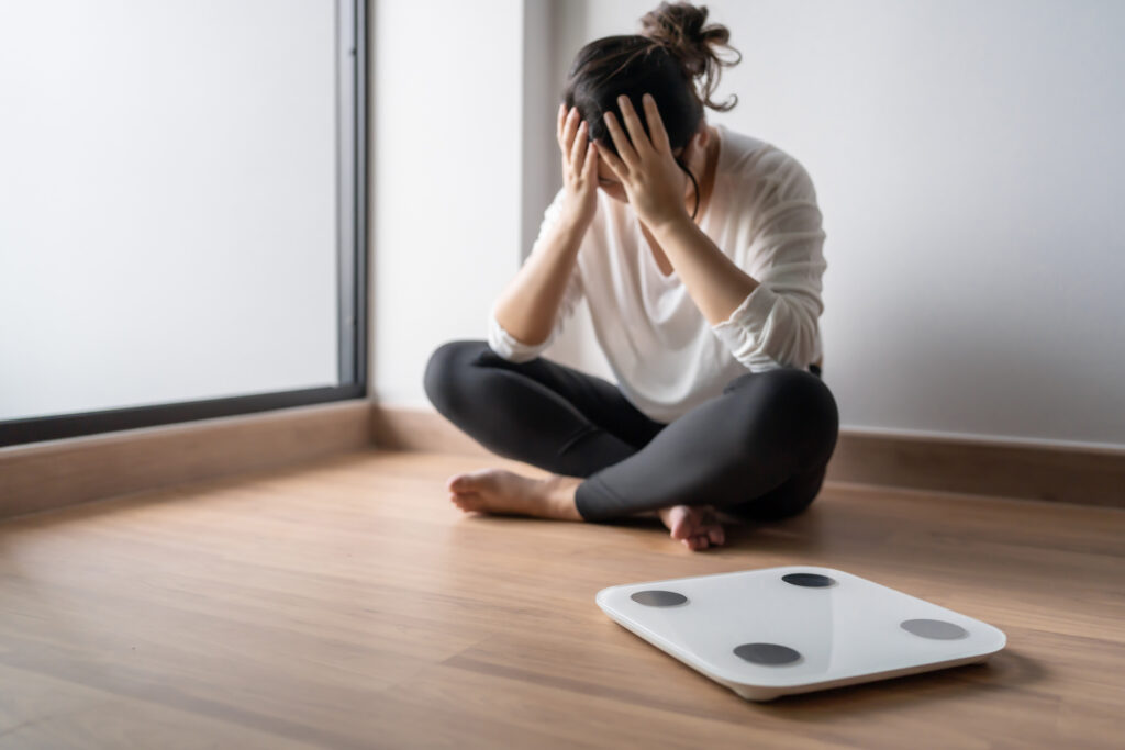 A woman sits cross legged on the floor beside a scale, holding her head in her hands, concerned with menopause weight gain. 