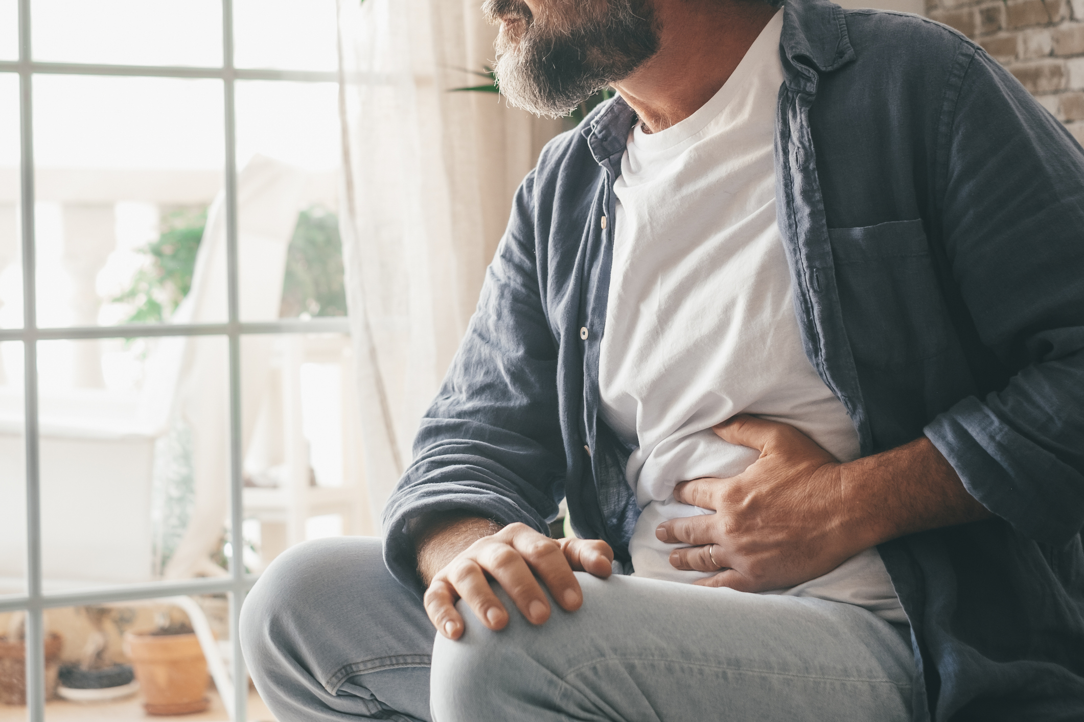A man is sitting down by a window, holding his lower belly in discomfort.