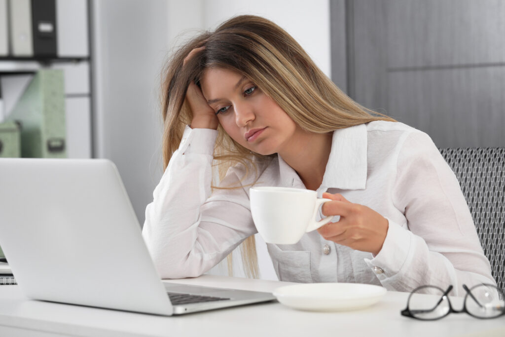 A sluggish young woman sits in front of her laptop, a coffee cup in one hand and holding her head with the other.