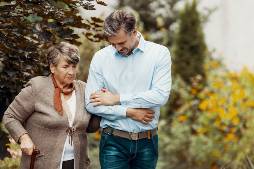 An adult male escorts his elderly mother on a walk in the garden. 