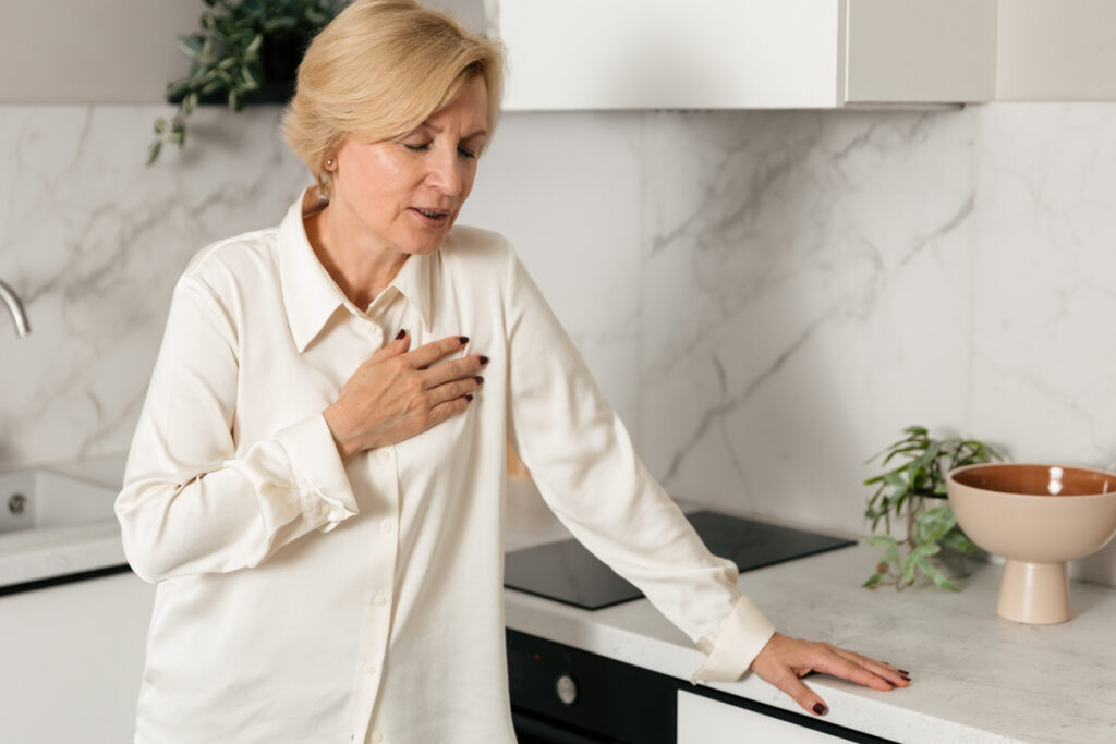 An older woman is holding her chest in pain in the kitchen.