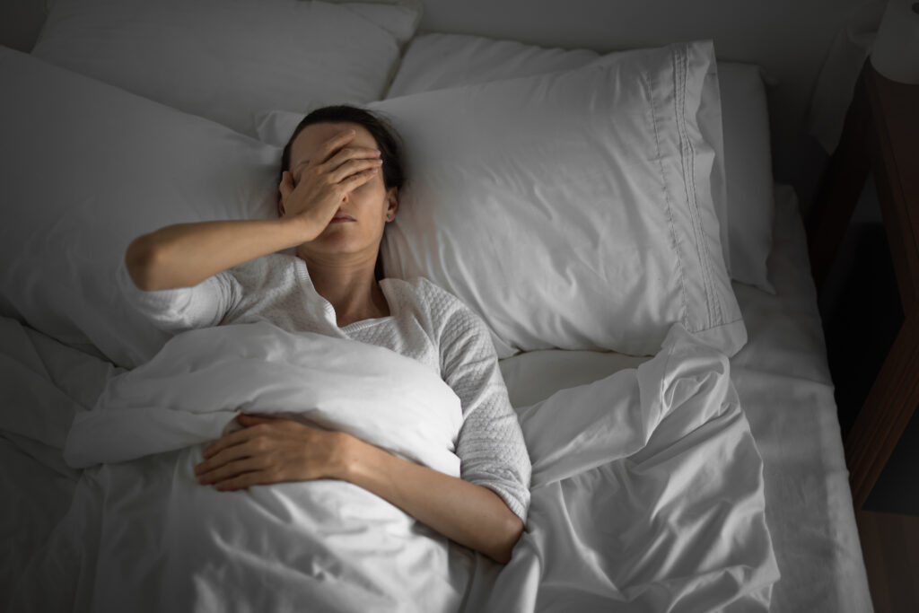 A woman is lying in bed, holding her face in frustration due to sleeplessness, a factor in menopause weight gain. 