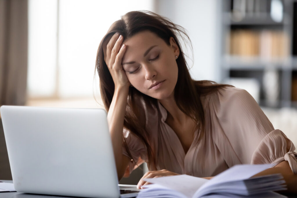 A tired young woman sits in front of her laptop, holding her head in her hand. 
