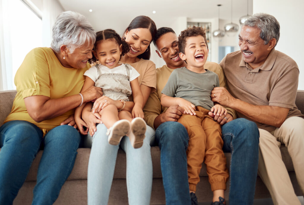 A multi-generational family sit laughing, side-by-side on a sofa. 