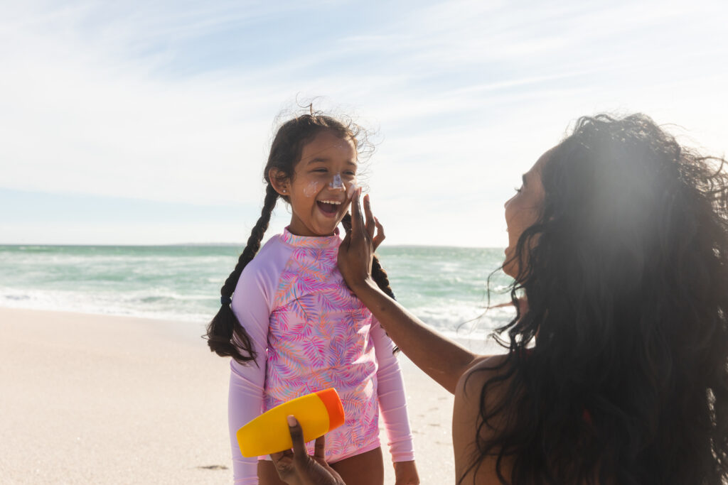 A mom applies sunscreen to her young daughter's face at the beach. 