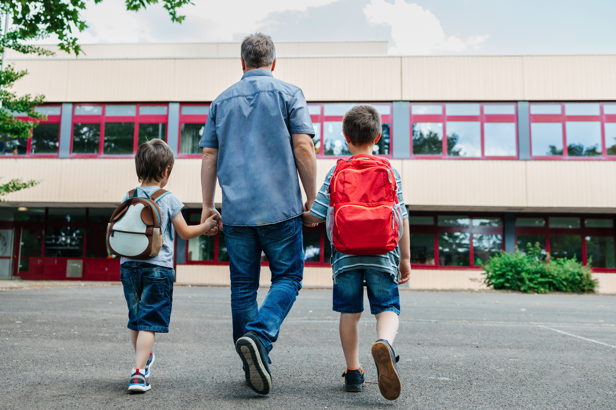 A father holds his two son's hands as they walk towards the school.