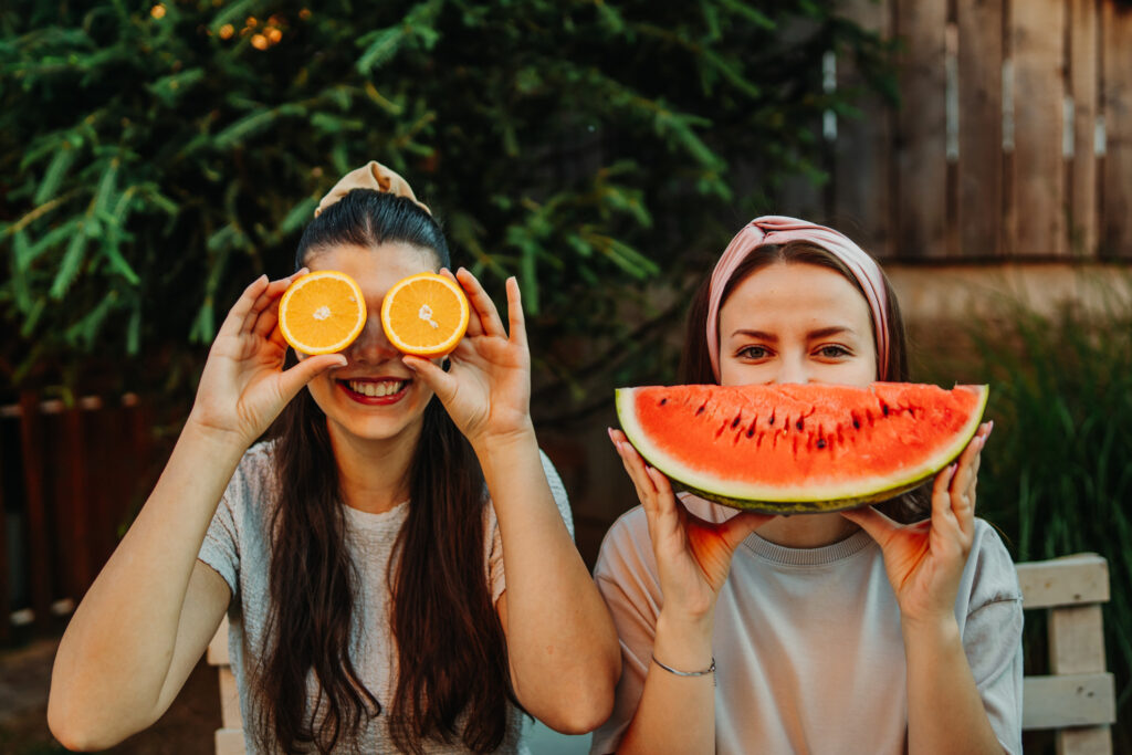 Two smiling young women playfully covering their faces with orange and watermelon.