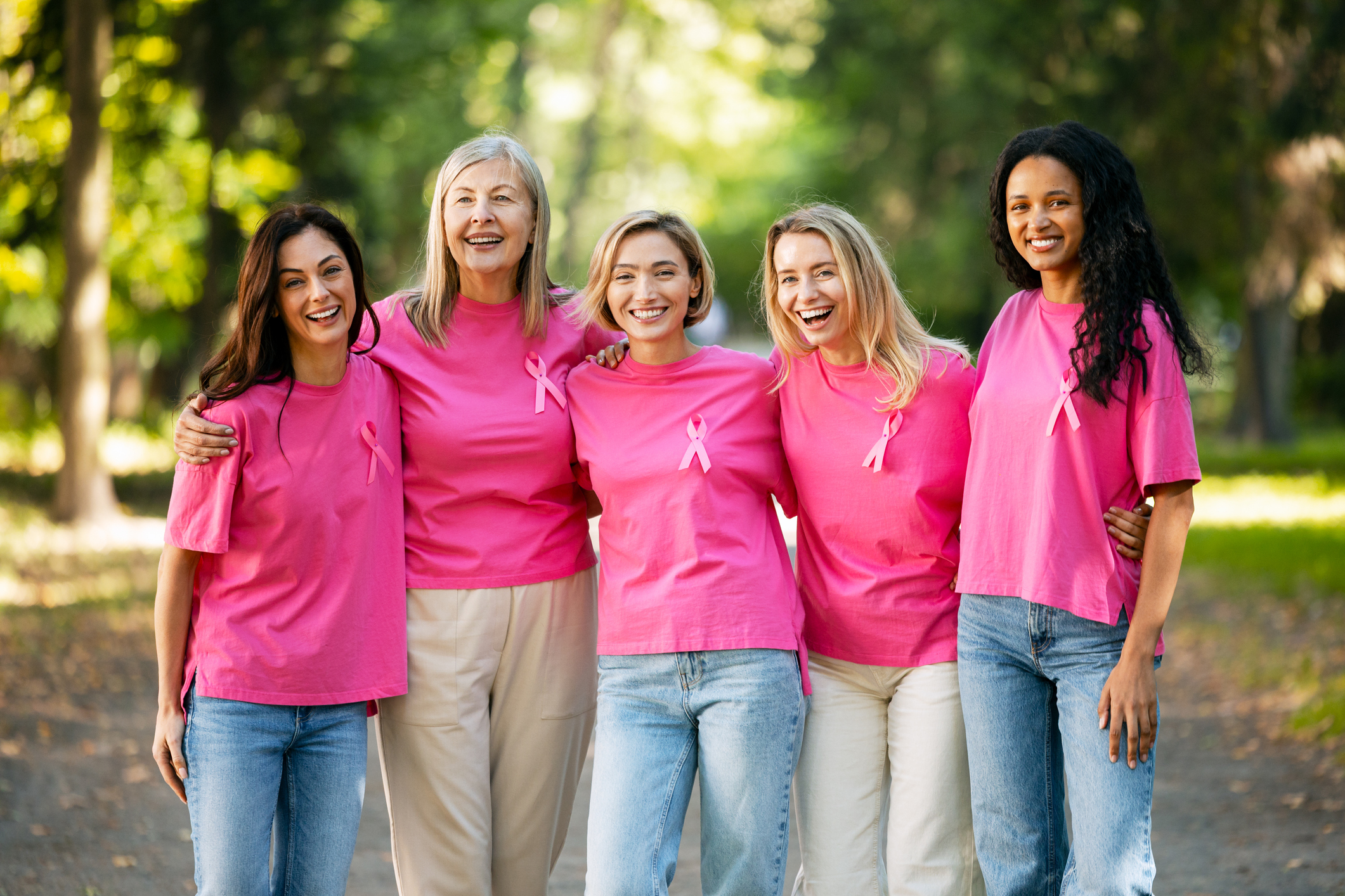 A diverse group of women wear pink t-shirts with Breast Cancer Awareness ribbons, smiling in the park.