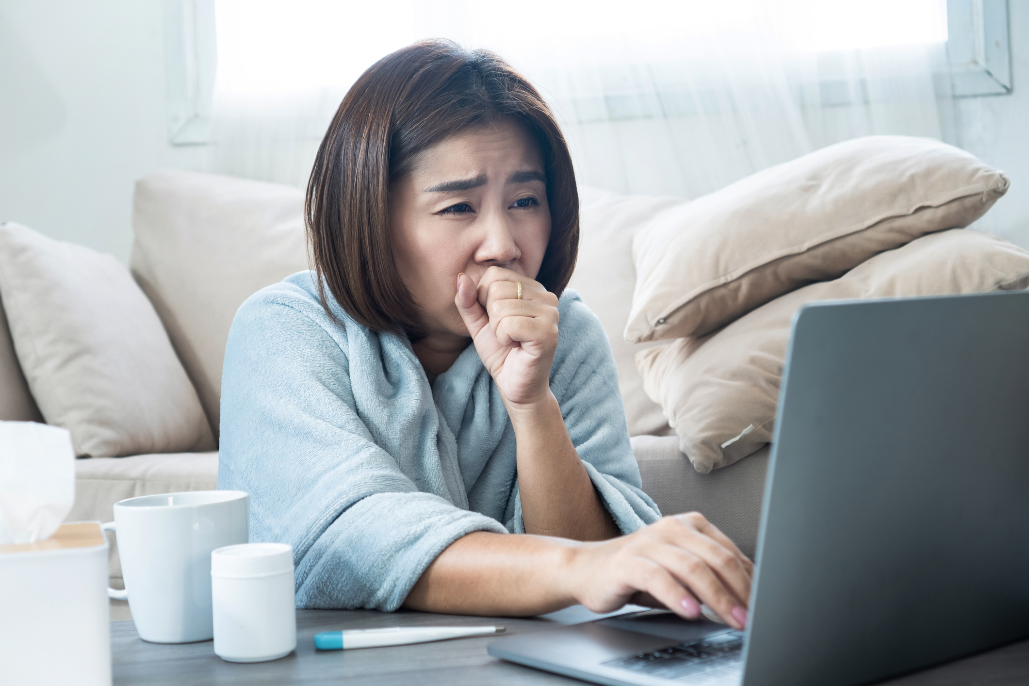 An unwell woman coughs while researching on her laptop in her living room.