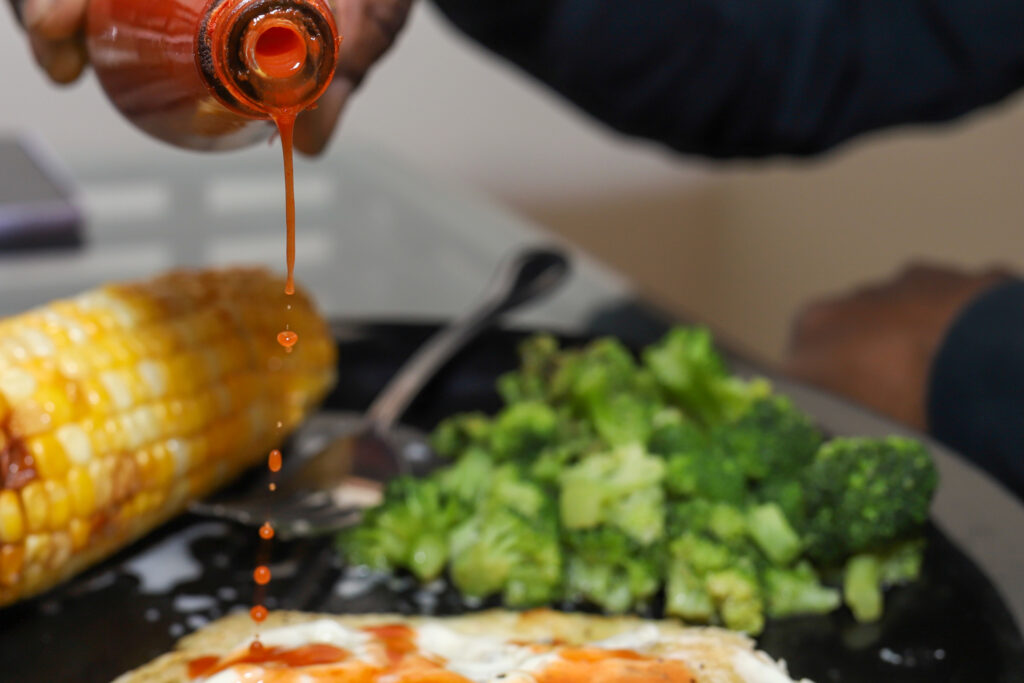 Hot sauce is being poured on a plate of eggs, corn on the cob and broccoli.