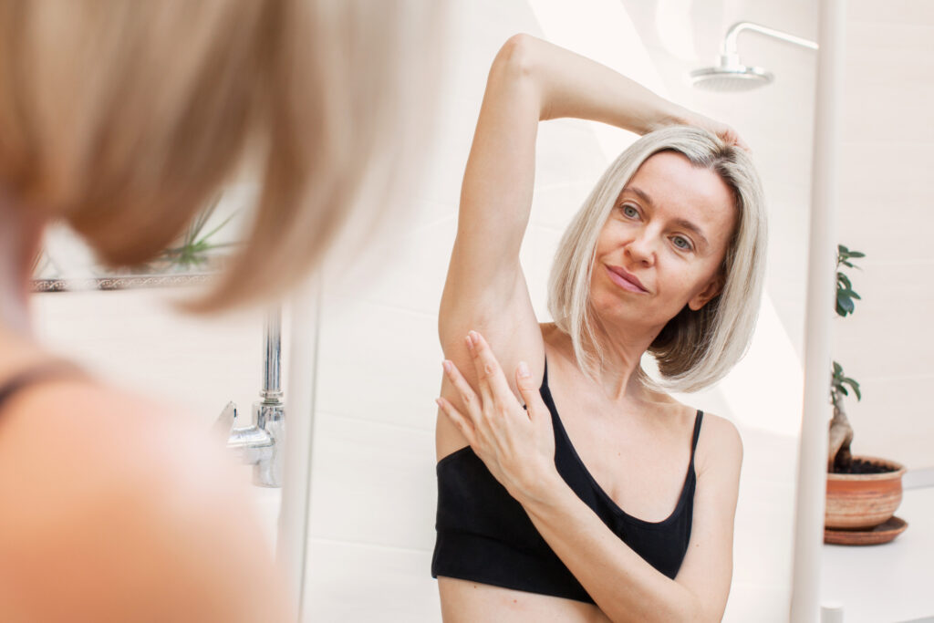 A middle aged woman is looking  in a mirror while doing a breast self-examination.